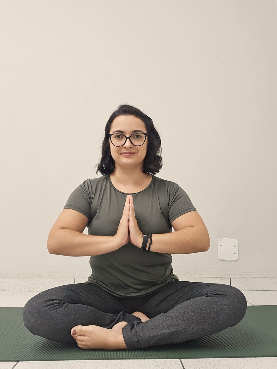 Bruna sentada em meditação sobre o tapetinho, com as mãos unidas em frente ao peito
